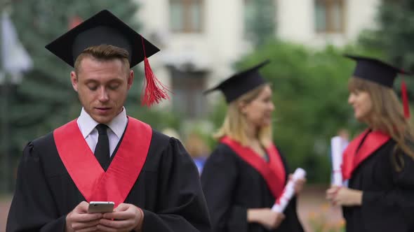 Man in Academic Dress and Cap Browsing on Phone, Women Chatting on Background