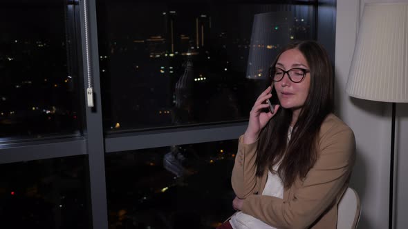 Woman Talking Phone Sitting Near the Window with Panoramic City View at Night