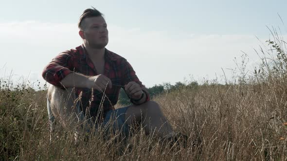 A Pensive Young Man In Nature Sits In Tall Grass And Looks Around. Sad Man In The Field.