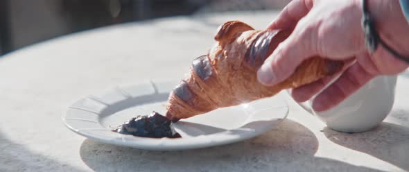 Hands of a man grabbing croissant and dip it in jam from a sunny balcony table closeup