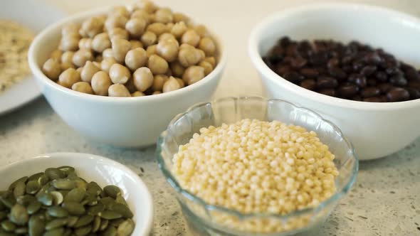 Various legumes, seeds and grains in bowls on white bench, pan close up