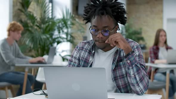 Focused Young African American Man Surfing Internet on Laptop with Blurred Colleagues Working Online