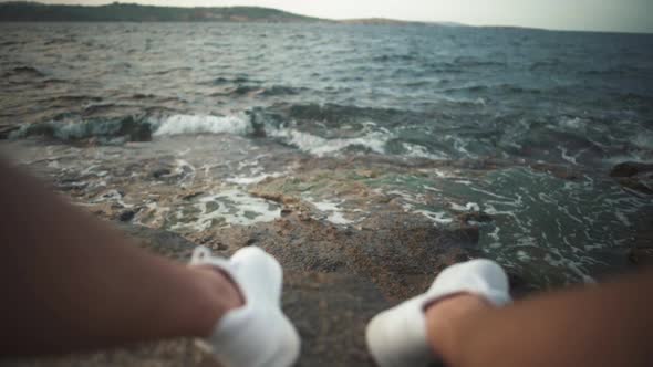 Legs in White Sneakers on a Rocky Beach on the Background of Sea Waves