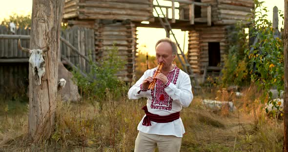 Man in a Folk SlavicUkrainian Costume Plays a Double Pipe in a Village Courtyard