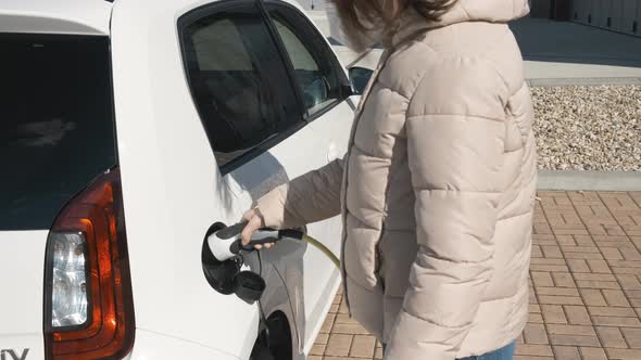 Woman in Protective Face Mask or Respirator Plugging an Electric Car for Charging in the Street at