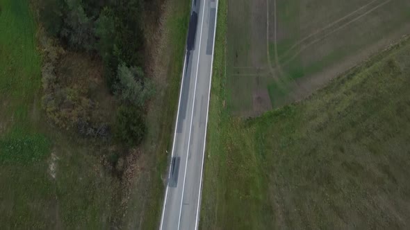 Aerial top shot of a small country road with green meadow surrounding it. A car passes by.
