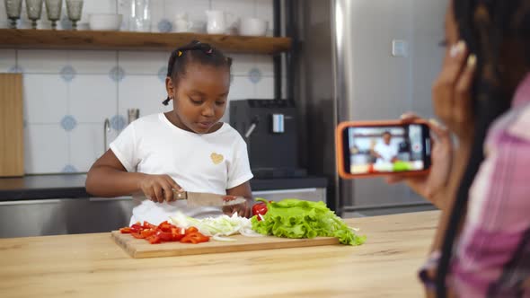 African Mother with Smartphone Recording Video of Cute Little Daughter Cooking Salad in Kitchen