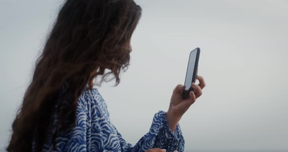 A Young Woman with Long Curly Hair Takes a Photo of the Sea on a Smartphone