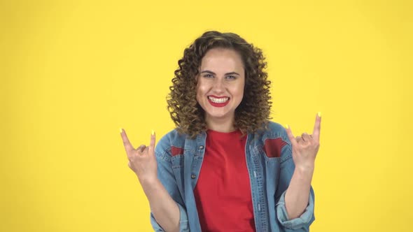 Curly Female Making a Rock Gesture, Enjoying Life and Laughing on Yellow Background at Studio, Slow