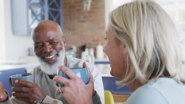 Mixed race senior couple holding coffee cups smiling looking at each other at home