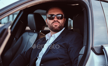Businessman in sunglasses dressed in a formal suit sitting on the front seats in the luxury car.