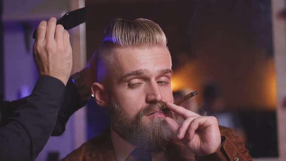 Young Caucasian Barber Cutting Hair of His Cool Rich Client Sitting in Armchair and Smoking Cigar.