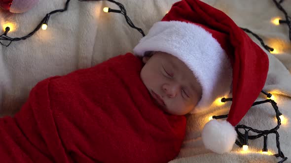 Top View Portrait First Days Of Life Newborn Cute Funny Sleeping Baby In Santa Hat Wrapped In Red