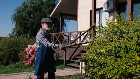 Retired Grandfather Takes Care of Plants Watering Trees in the Garden Near the House