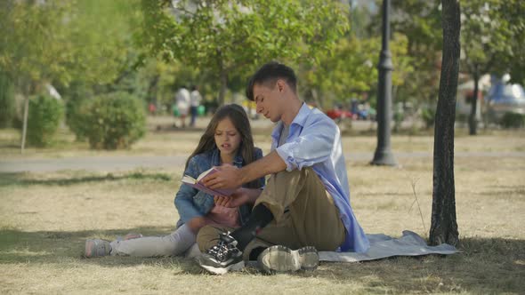 Teenage Boy with His Small Sister Reading Book with Poems or Tales and Discussing Sitting Outdoor in