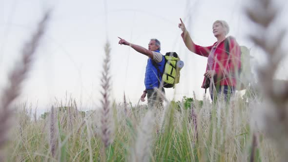 Senior hiker couple with backpacks pointing towards a direction while standing in the grass field.