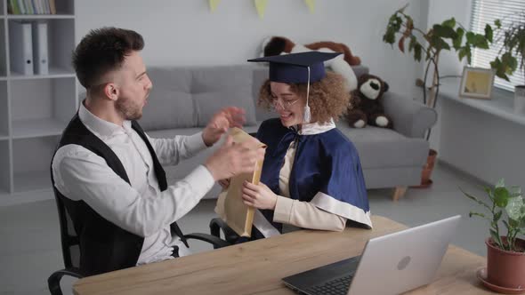 Remote Education University Graduate in Academic Dress Receives Diploma From Her Husband During an