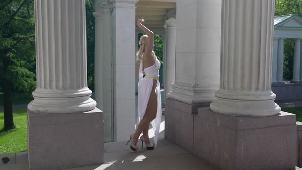 a Blonde in a White Toga and Highheeled Shoes is Dancing Among the Columns