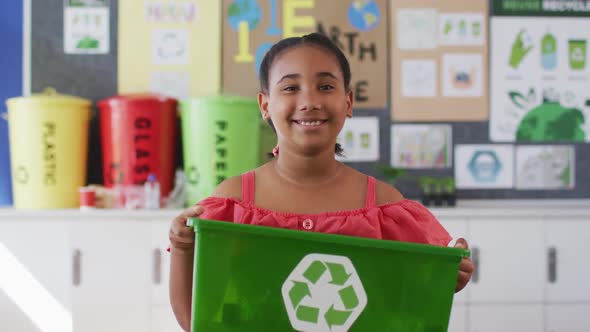 Mixed race schoolgirl smiling, holding recycling bin, standing in classroom