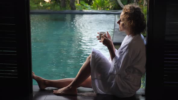 Side View of Woman Wearing Bathrobe Enjoying Her Morning Coffee Sitting on the Edge of the Swimming