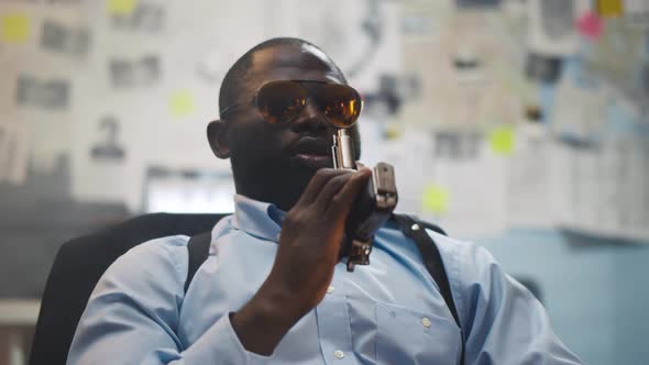 Confident African Policeman in Glasses Sitting at Desk with Gun at Police Station