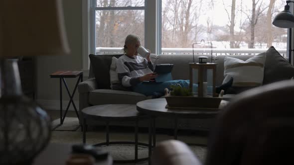 Woman sitting on the couch in front of a bay window, drinking coffee while scrolling on a tablet fro