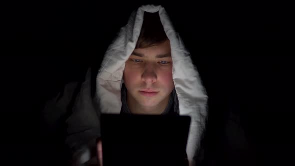 A Young Man Lies Under a Blanket with a Tablet in Him Hands. A Man in the Dark Uses a Pad. Modern