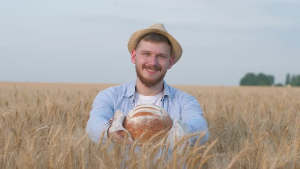 Young Happy Grain Grower, Handsome Man in Straw Hat Gives You at Camera Tasty Baked Bread and Smiles