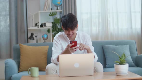 Asian Man Touching Smartphone And Laughing While Using Laptop Computer In The Living Room