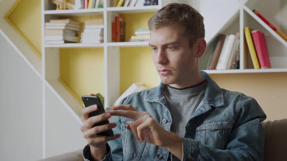 Smiling Young Man Holds Mobile Phone While Sitting on Sofa at Home and Celebrating