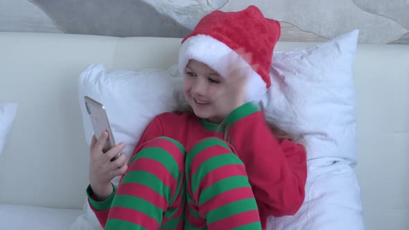Happy Little Girl in Santa Hat and Red Pajama Enjoy a Video Phone Call with Relatives or Friends on alt