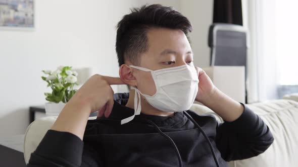 A Young Asian Man Puts on a Face Mask and Looks Seriously at the Camera As He Sits on Couch at Home