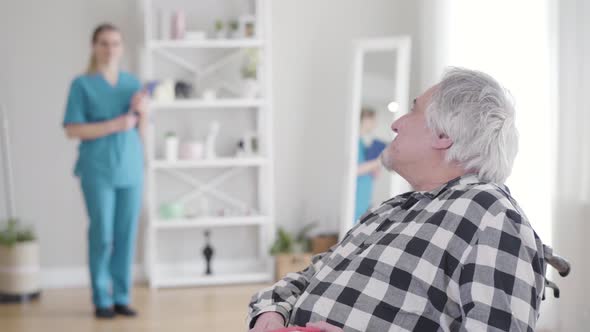 Portrait of Happy Caucasian Grey-haired Retiree Looking Back at Nurse, Turning To Camera and