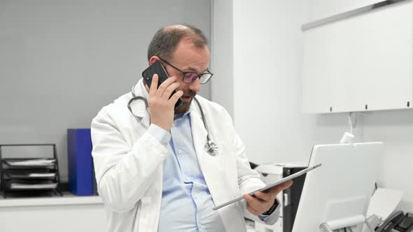 Male Physician with Stethoscope Sitting at Desk Talking on Mobile Phone and Looking at Digital