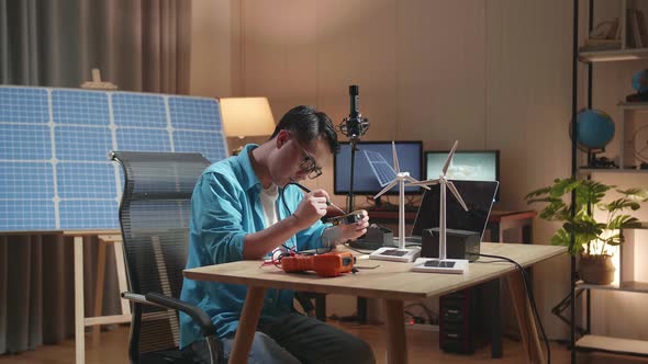 Asian Man With Wind Turbine Fix The Circuit Board While Working With Laptop Next To The Solar Cell