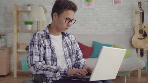 Young Asian Man in Stylish Glasses Disabled in a Wheelchair with a Laptop in the Living Room of the