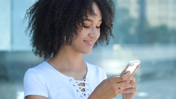 Lovely African American Teen Girl Typing Message To Friend on Phone, Clicks on Display, Surfing