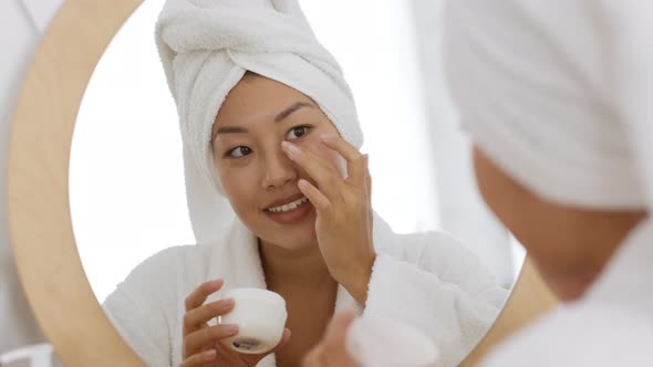 Young Carefree Korean Woman Applying Moisturizing Cream on Face Doing Beauty Care Procedures After