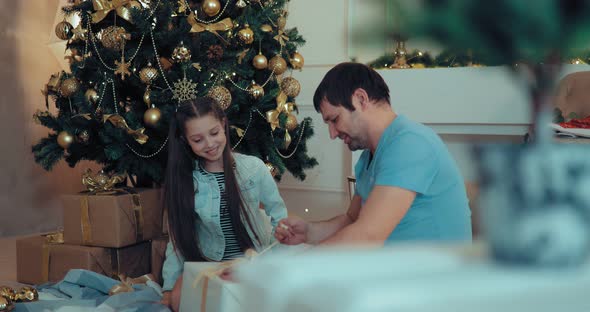 Girl and Dad Sit on a New Year Tree and Open Boxes of Presents