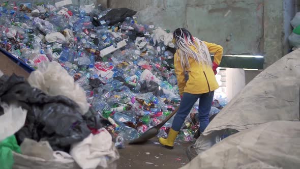 A Girl with Dreadlocks in Yellow Boots Scoops Used Bottles with a Large Duck Shovel at a Plastic