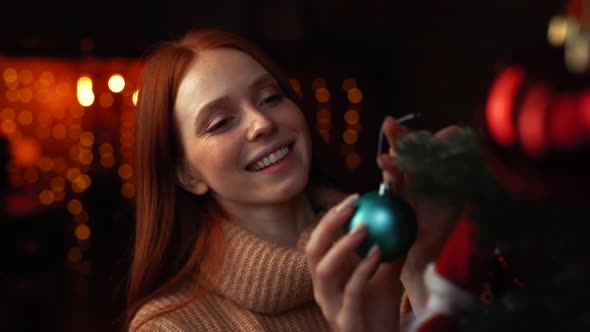 Close-up of Smiling Redhead Young Woman Hanging an Christmas Ornament on Xmas Tree at Cozy Room