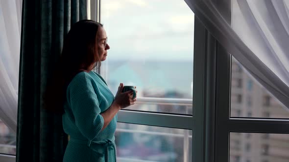 an Attractive Woman at Home in the Morning in a Bathrobe Stands at the Window and Drinks a Hot Drink