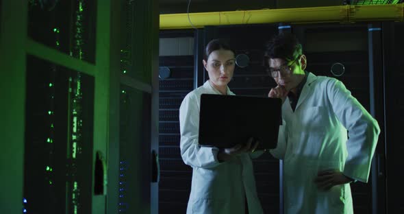 Diverse female and male it technicians in lab coats using laptop checking computer server