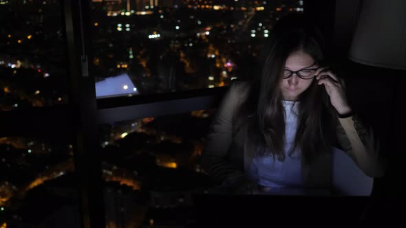 Tired Sleepy Woman Is Working on Her Computer at Night Near the Window with Cityscape