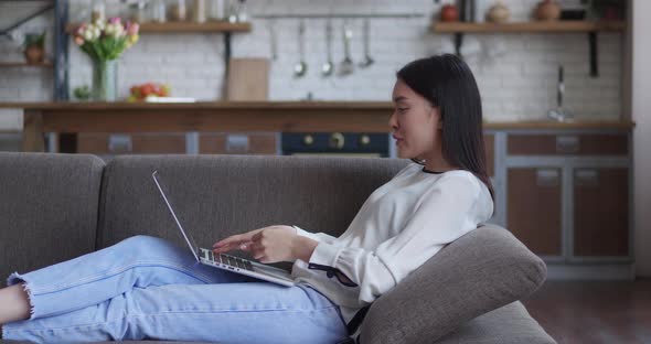 Asian Woman Using Laptop for Video Chat on Couch Kitchen Background
