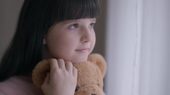 Cute Brunette Caucasian Girl with Brown Eyes Hugging Toy Looking Out the Window at Home