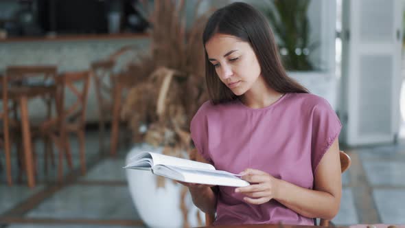 Portrait of Beautiful Woman Sitting at Table in Cafe and Reading Book