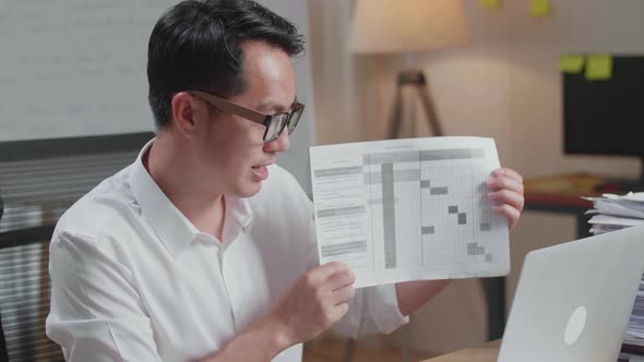 Asian Man With Documents Explaining Work On Paper While Having Video Call On Laptop