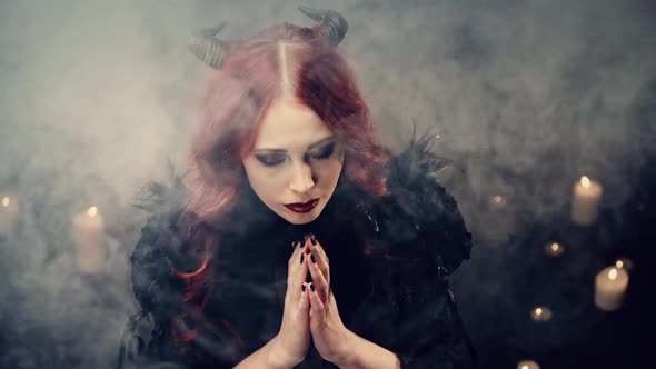 Red-haired Girl Witch with Black Horns Sitting on Floor in Candlelight and Praying with Folded Hands