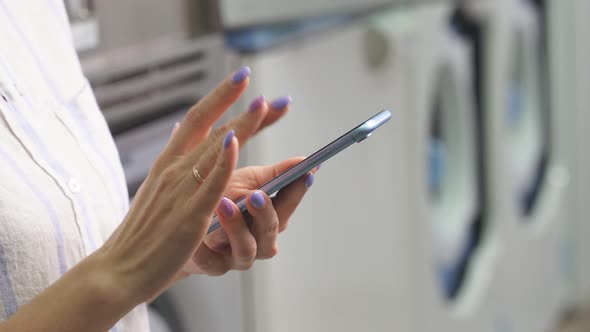 Closeup of a Woman's Hands with a Smartphone Against the Background of Washing Machines in a Public